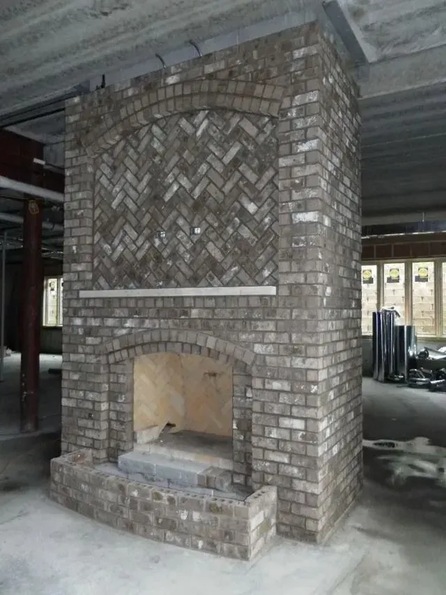 Brick fireplace under construction, interior setting. Gray bricks, arched opening, herringbone pattern detail.