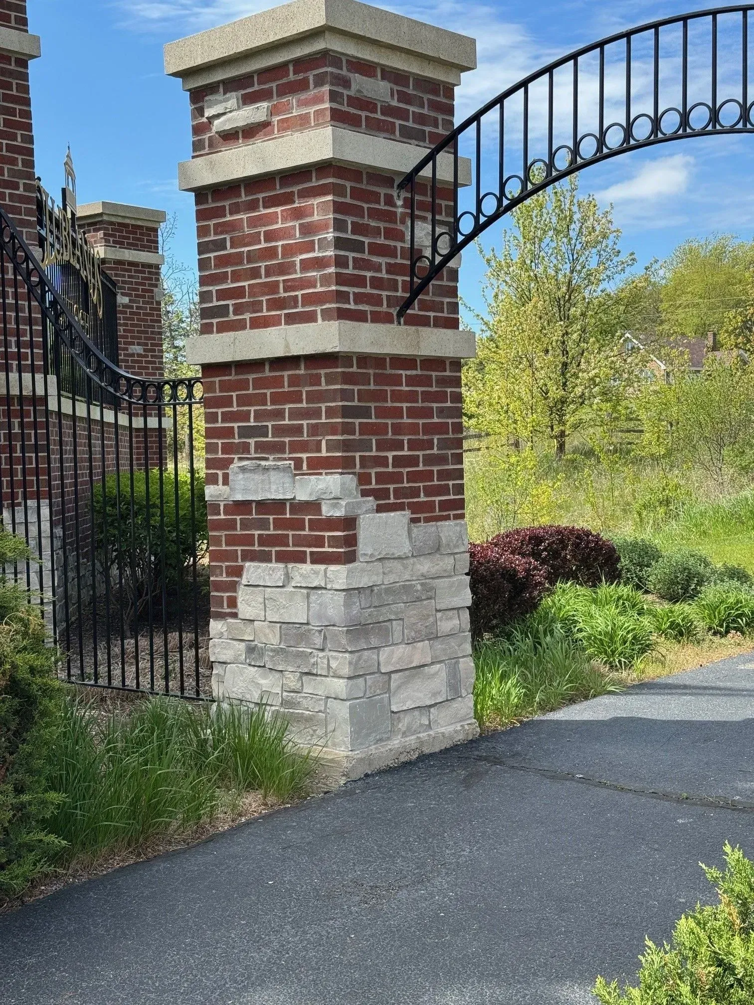 Brick and stone gate pillar with black metal arch, path, and greenery.