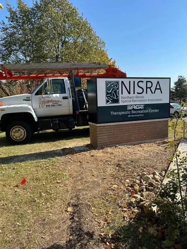 White truck with ladder beside a sign for NISRA, an association. Brown brick base, blue sky.