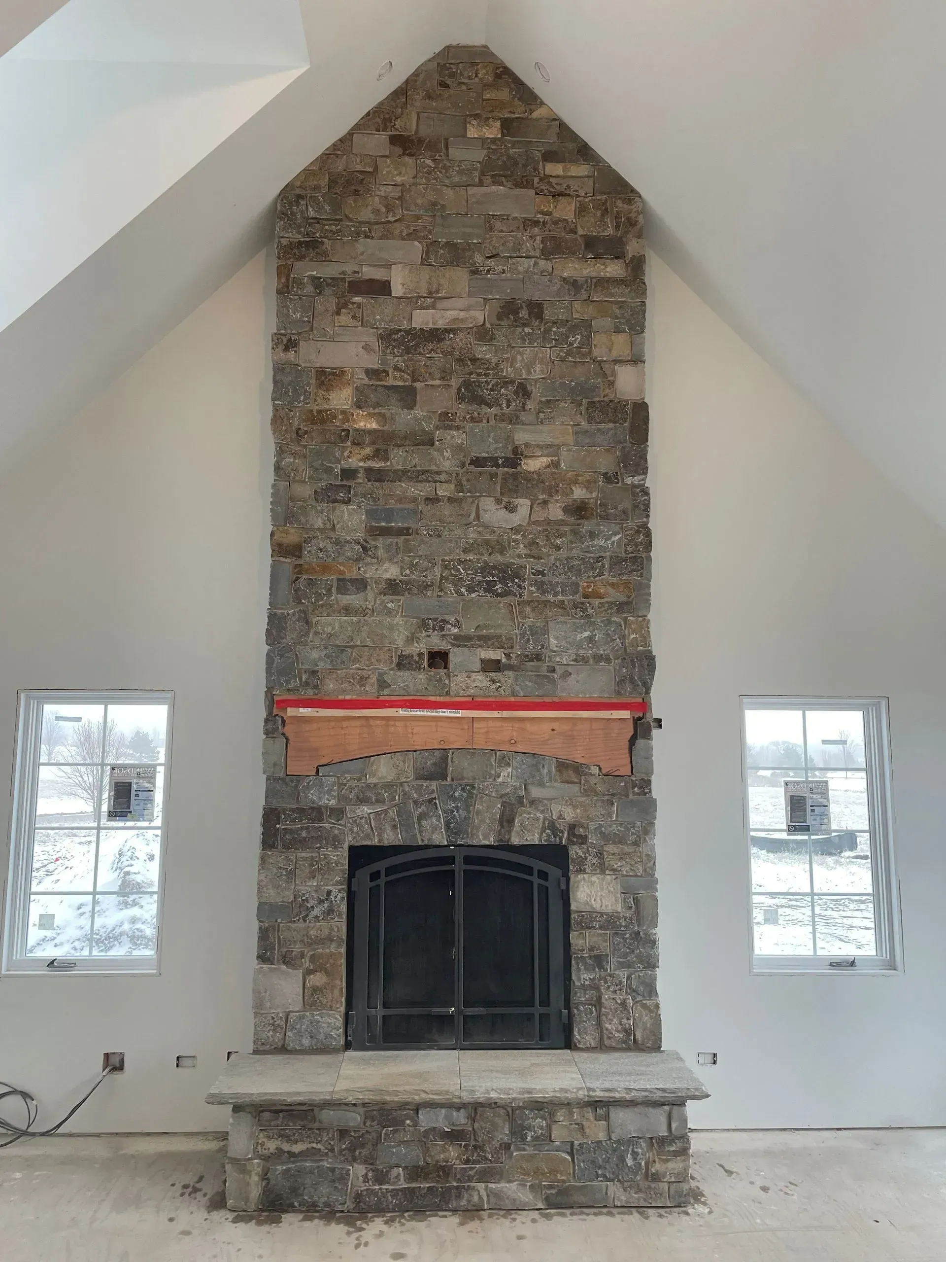 Stone fireplace in a white-walled room with two windows. The room has a high, slanted ceiling.