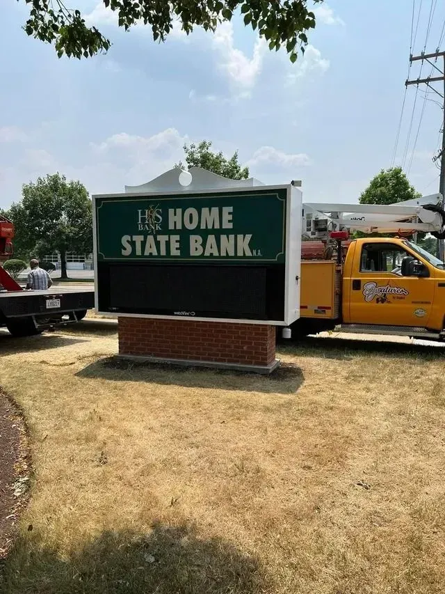 Home State Bank sign being worked on by utility trucks on a grassy area. The sign is dark green and white.