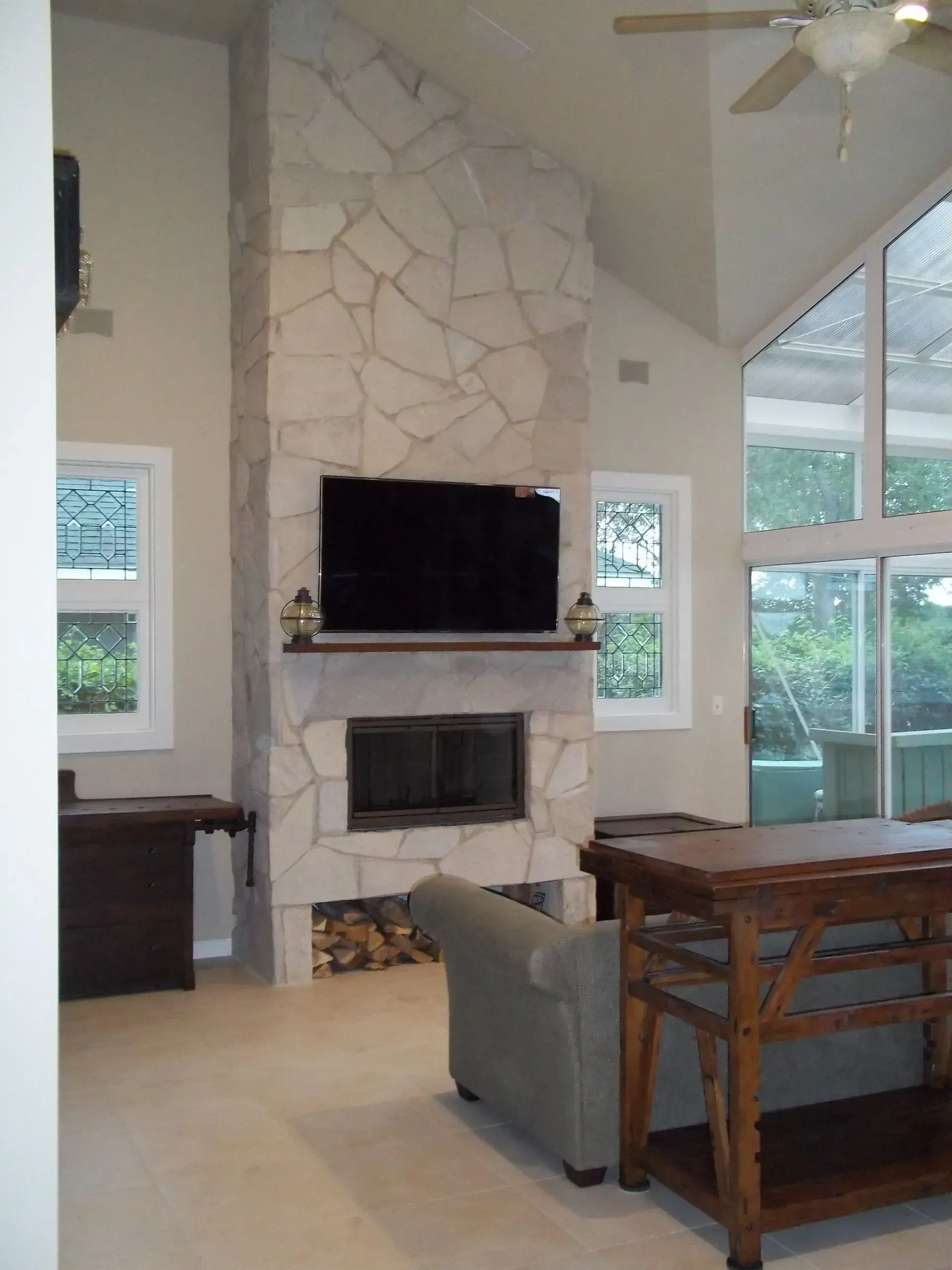 Living room with stone fireplace, TV, and large windows. Light walls, wood furniture, and gray sofa.