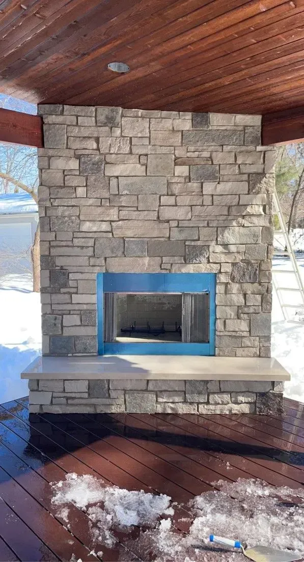 Stone outdoor fireplace with blue metal frame, on wooden deck, snow in background.