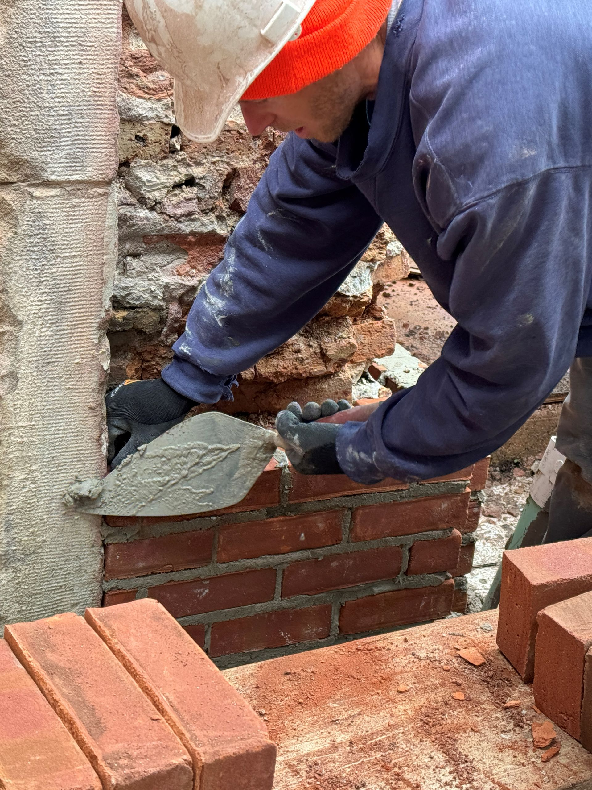 A construction worker laying bricks with a trowel, wearing gloves and a hard hat.