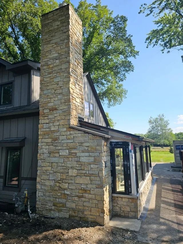 Stone chimney on a house with gray siding; black framed windows, blue sky.
