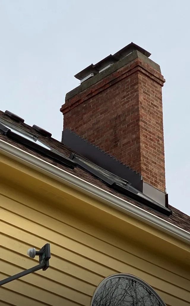 Brick chimney on a yellow-sided building roof, with metal flashing and cloudy sky.