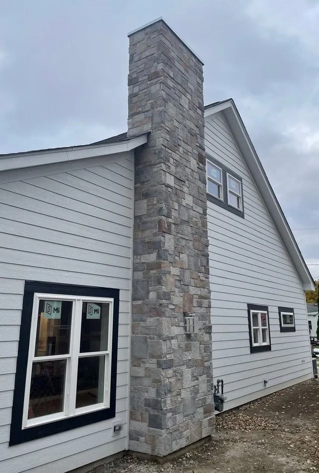 Stone chimney on a house with white siding and black window frames under an overcast sky.