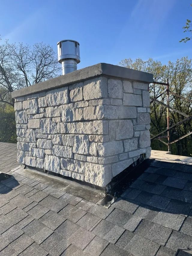 Stone chimney on a shingle roof with a metal flue. Bright sky and trees in the background.