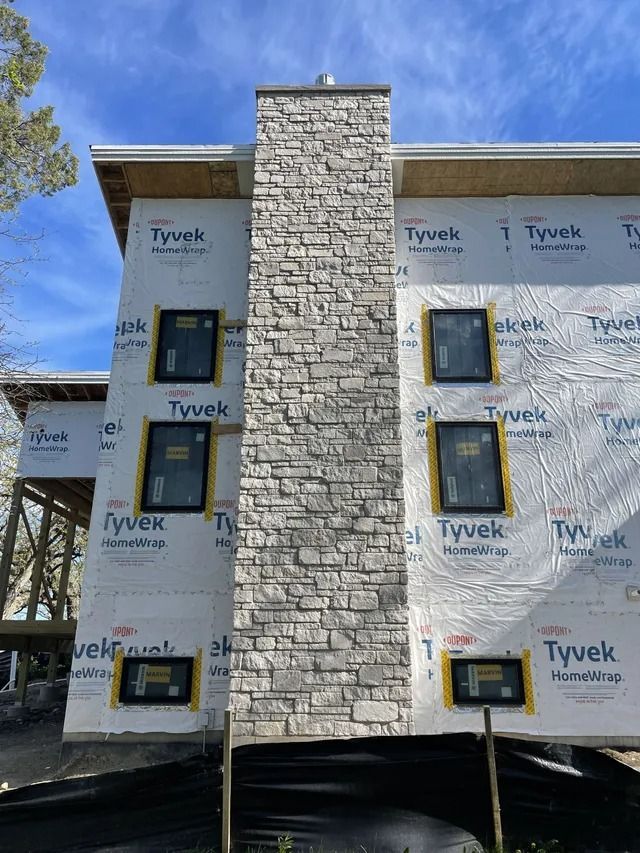 Exterior of a building under construction, featuring a stone chimney and windows covered in protective film.