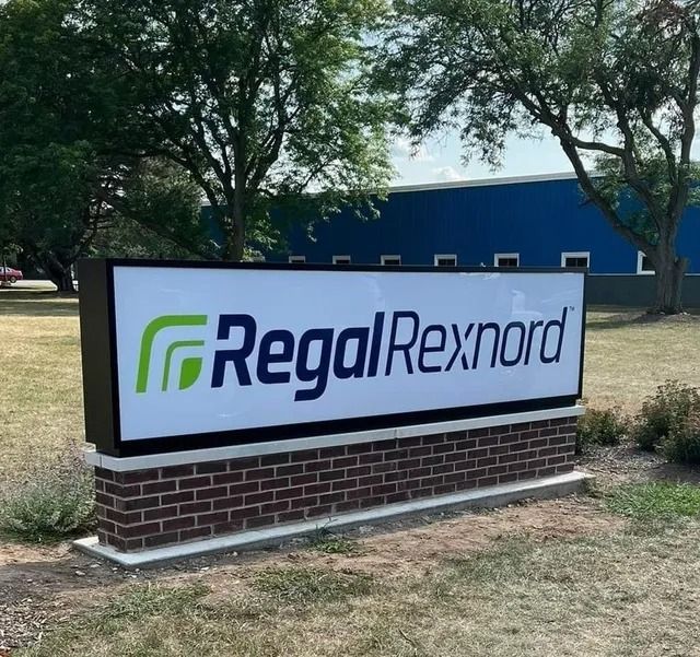 Sign for Regal Rexnord with green logo on white background, set on a brick base outdoors.