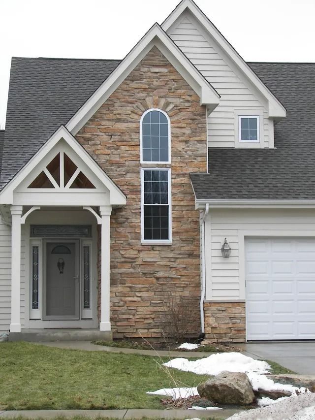Front porch with stone veneer, wreath on window, gray siding, concrete floor, flower garden.