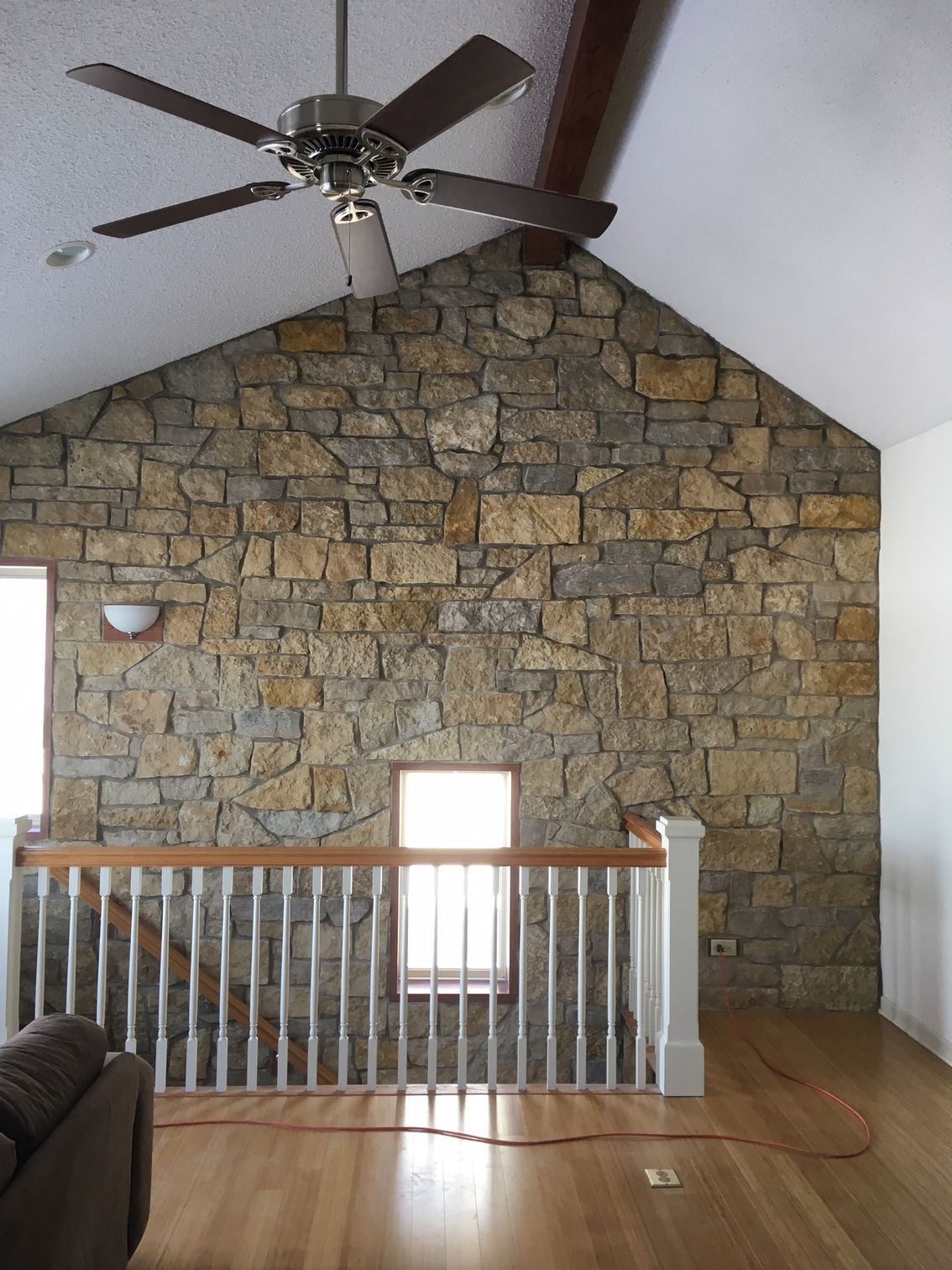 Stone wall with a wooden ceiling beam, a ceiling fan, and a wood floor. A staircase with a white railing is in front.