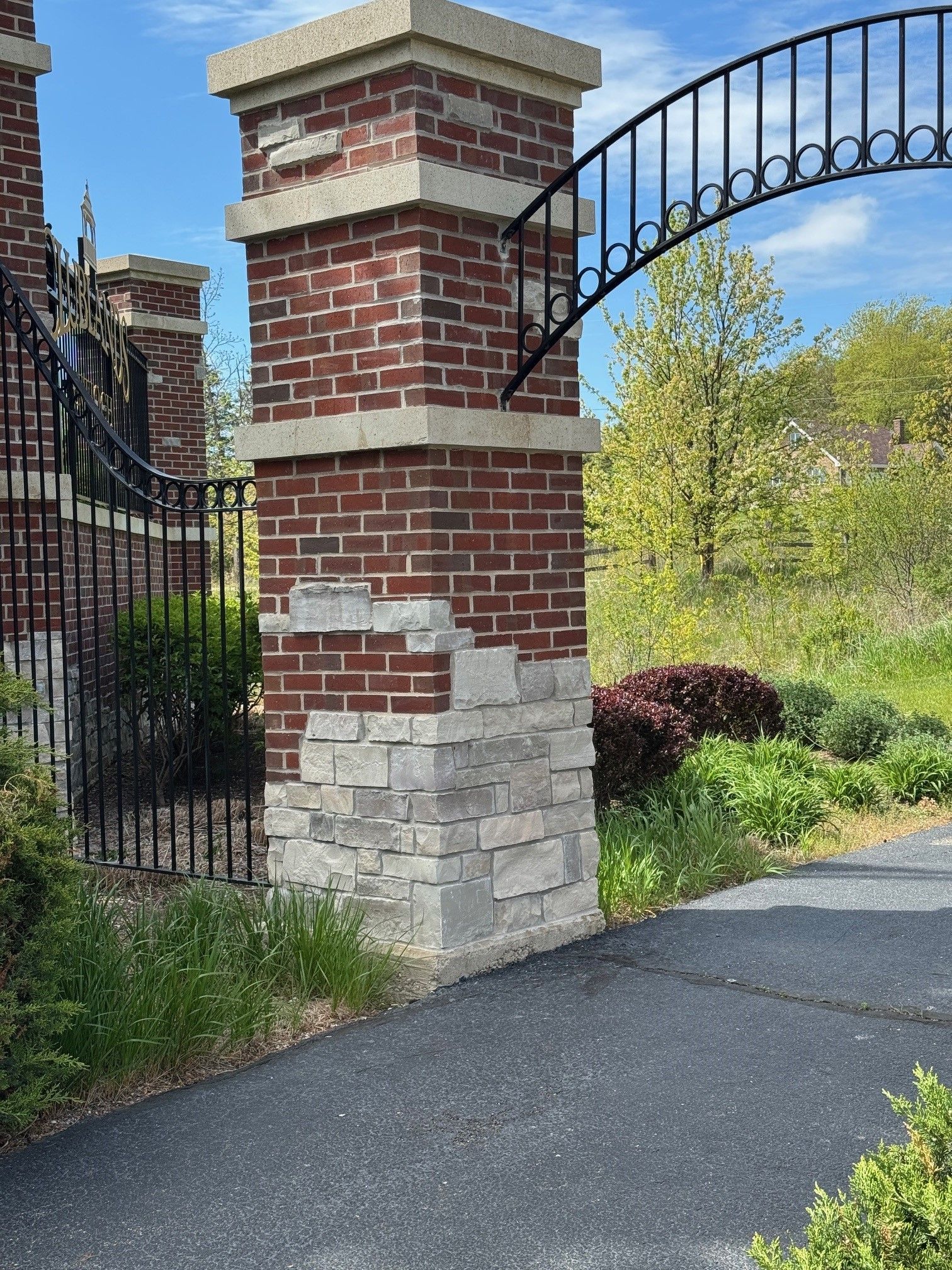 Brick and stone gate pillar with black metal archway, leading to a driveway with greenery.