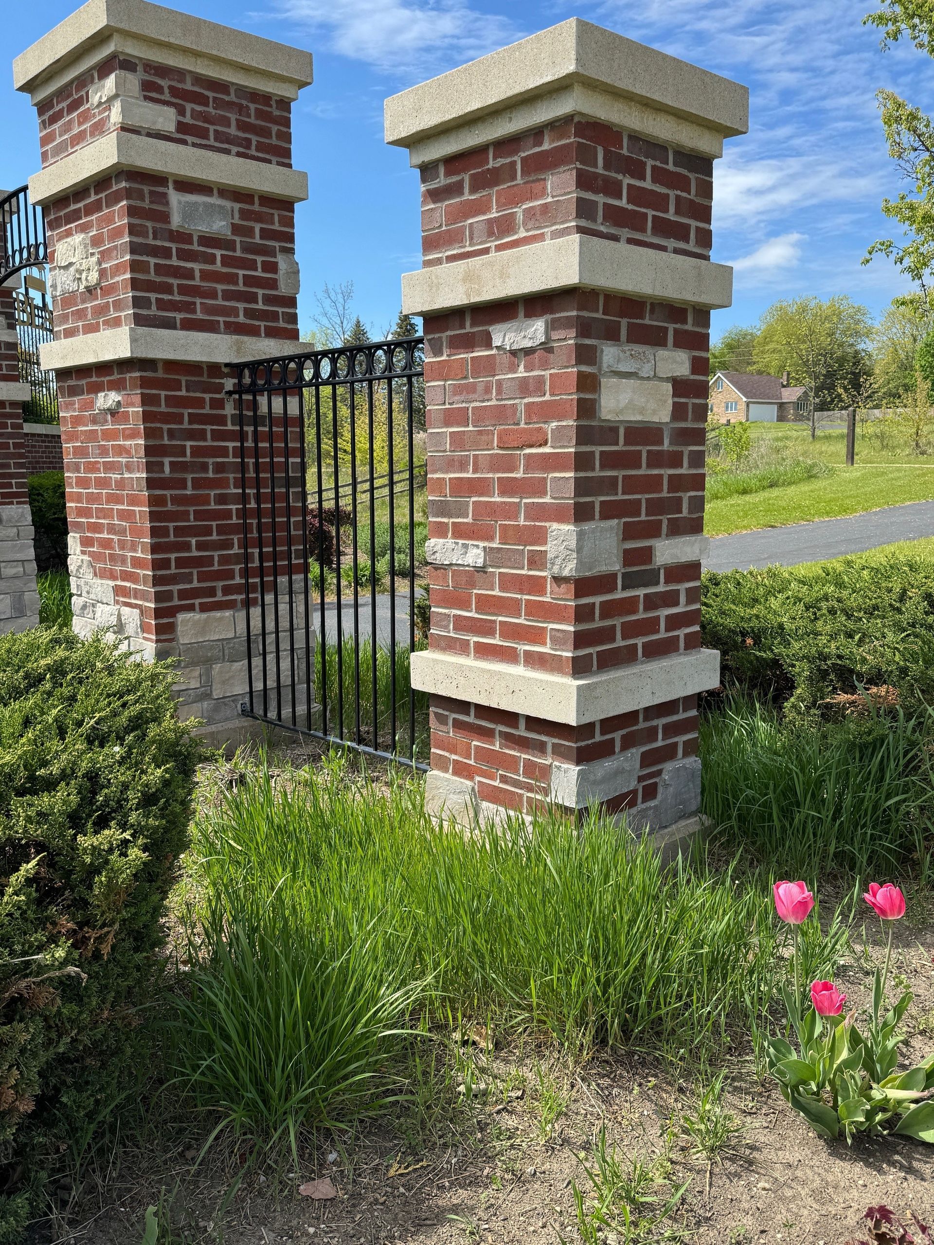 Brick pillars flank a black wrought iron gate, with greenery and pink tulips in the foreground.