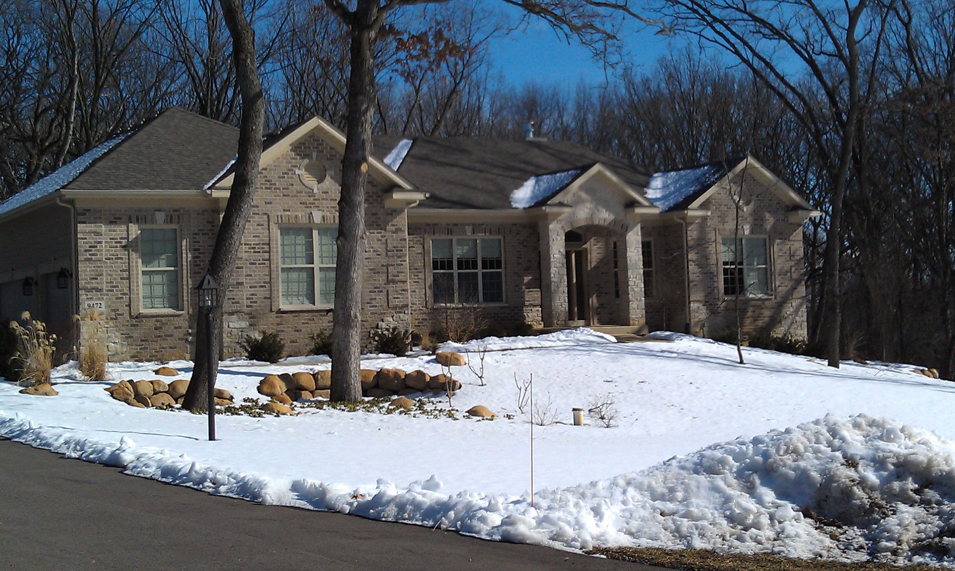 Stone-front house in a snowy setting with a dark roof and leafless trees under a bright blue sky.