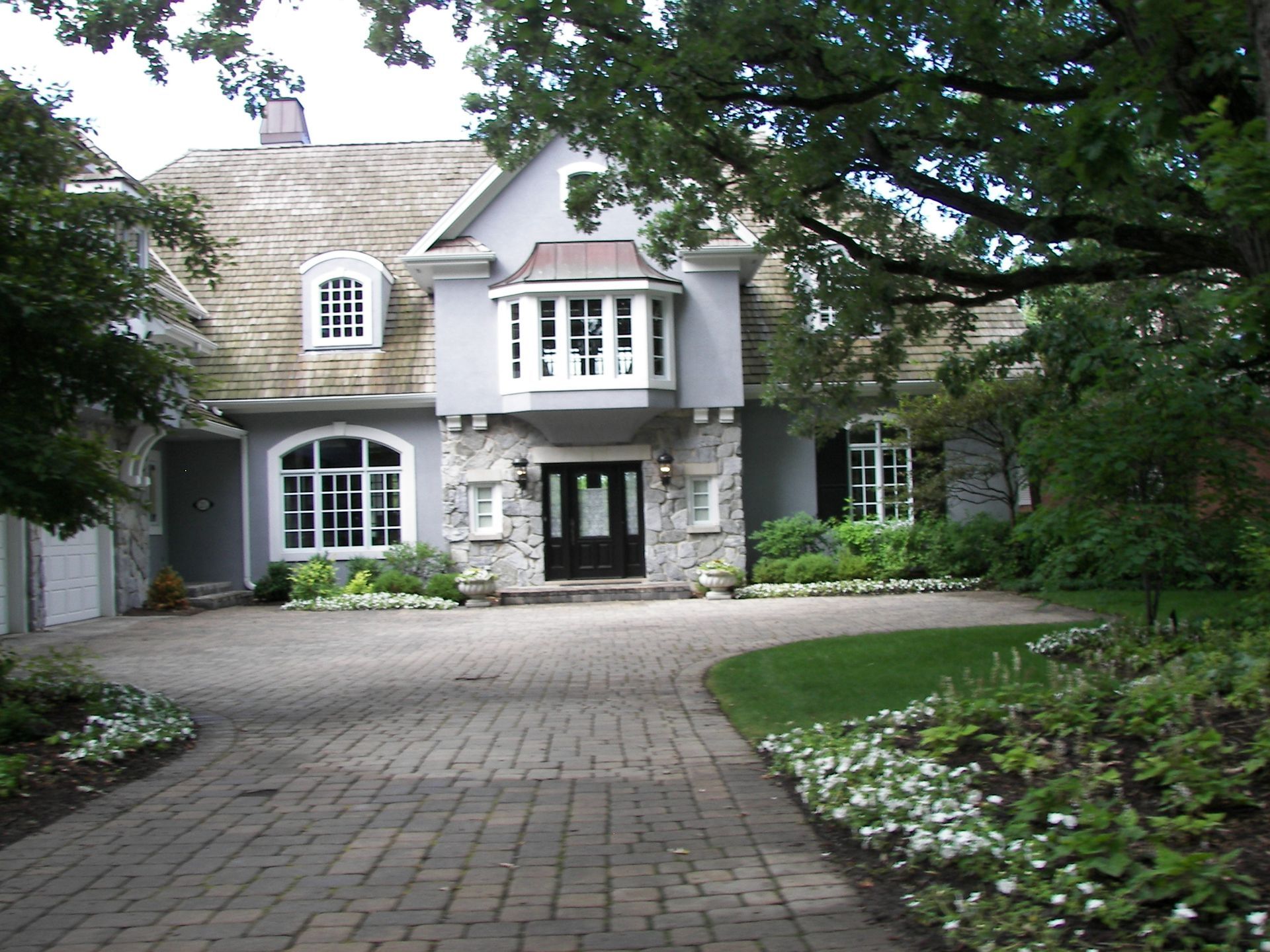 Elegant two-story house with stone facade, bay window, and cobblestone driveway lined with landscaping.