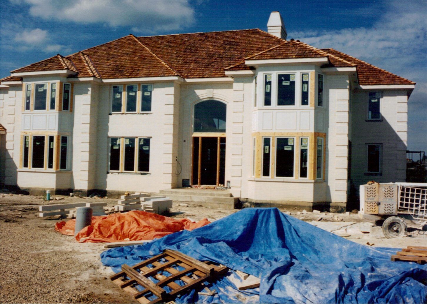 Two-story house under construction with light stucco exterior, brown roof, and blue tarp in the foreground.