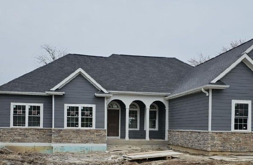 New house exterior under construction, gray siding and roof, stone base, white trim.