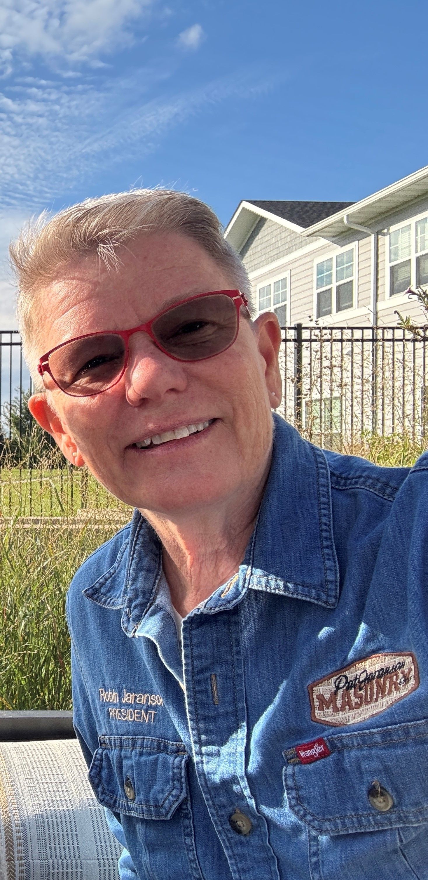 Person wearing sunglasses and denim jacket smiles at camera outdoors.