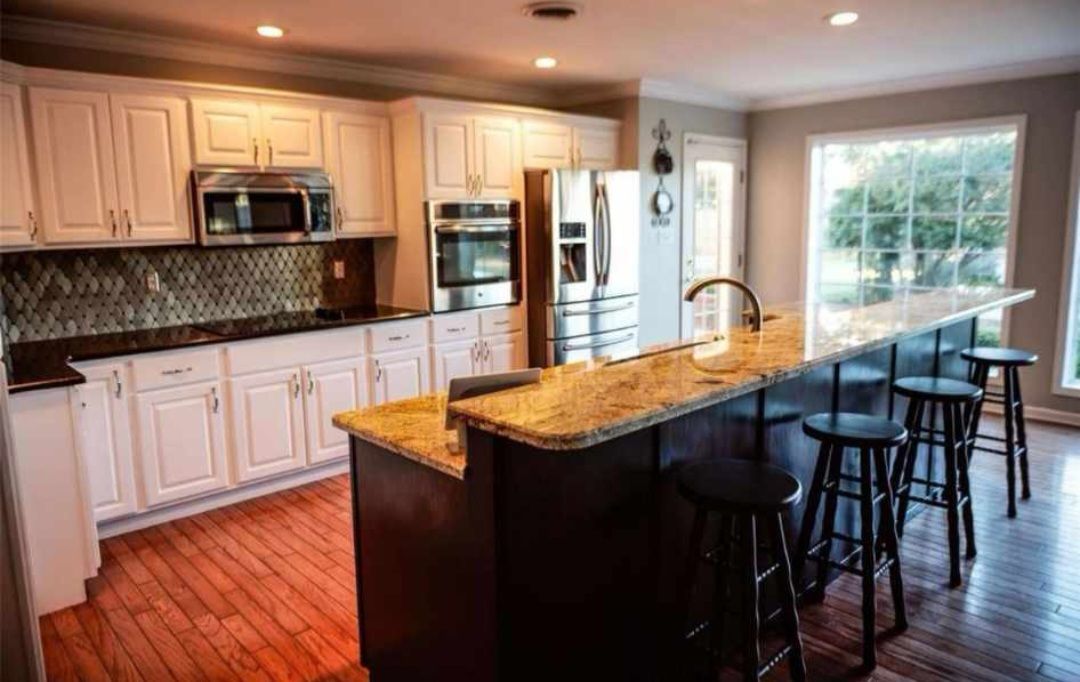 A kitchen with white cabinets , granite counter tops , stools and a large island.