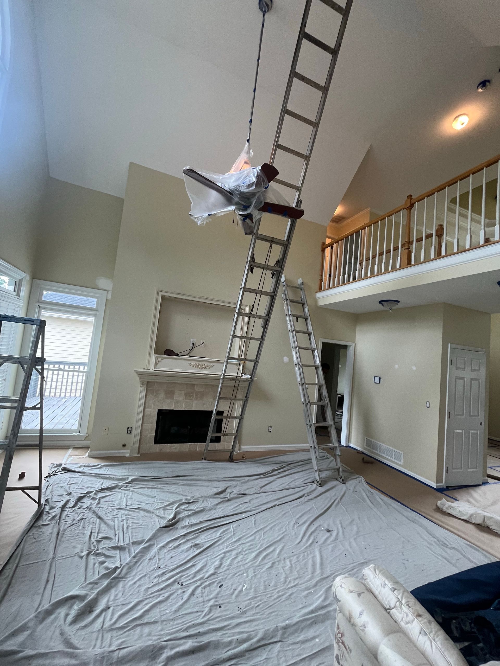 Man on ladder painting a high ceiling in a living room covered with drop cloths