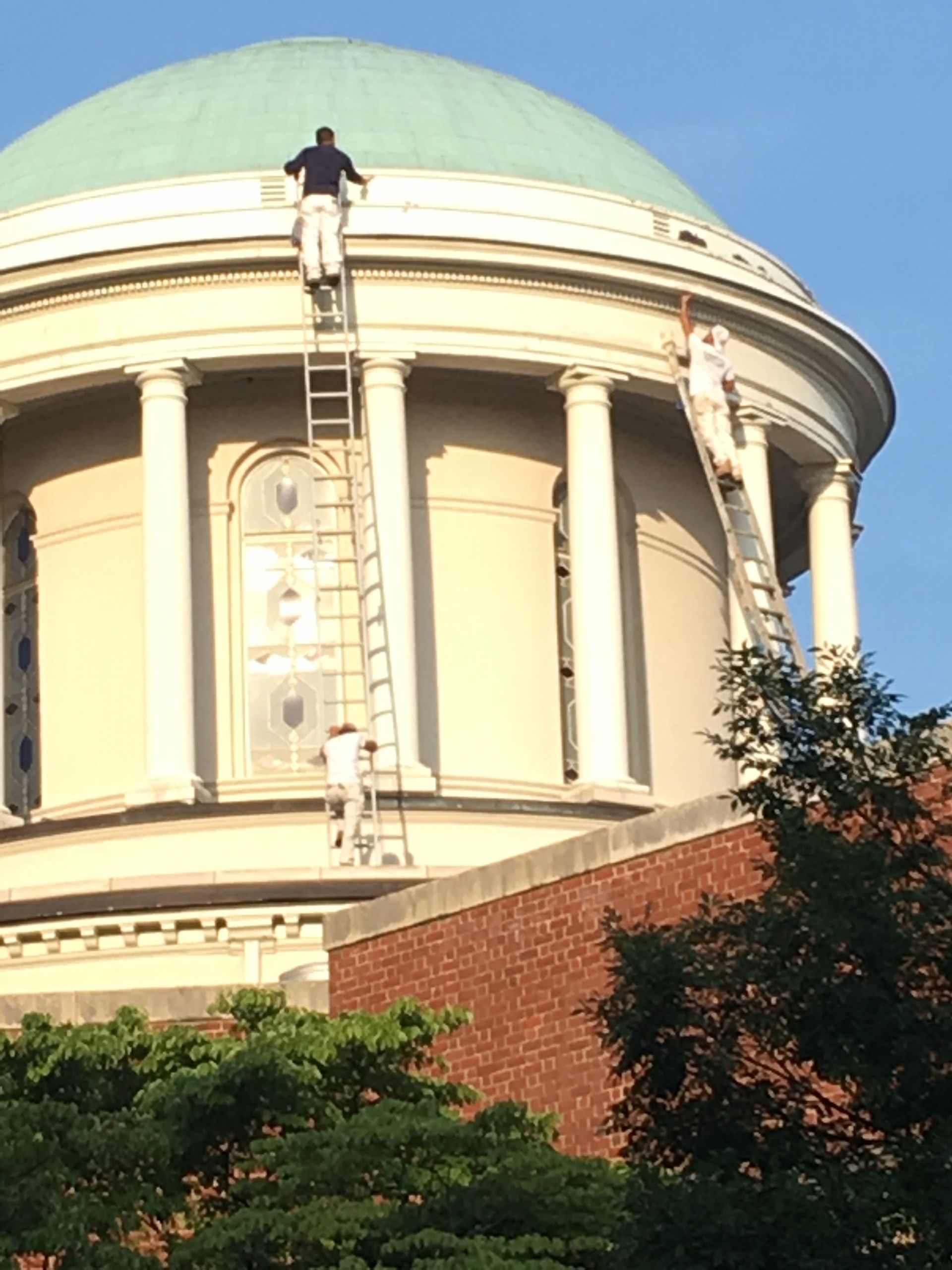 Historic building with a green dome and columns, one worker on the roof, framed by trees and brick walls