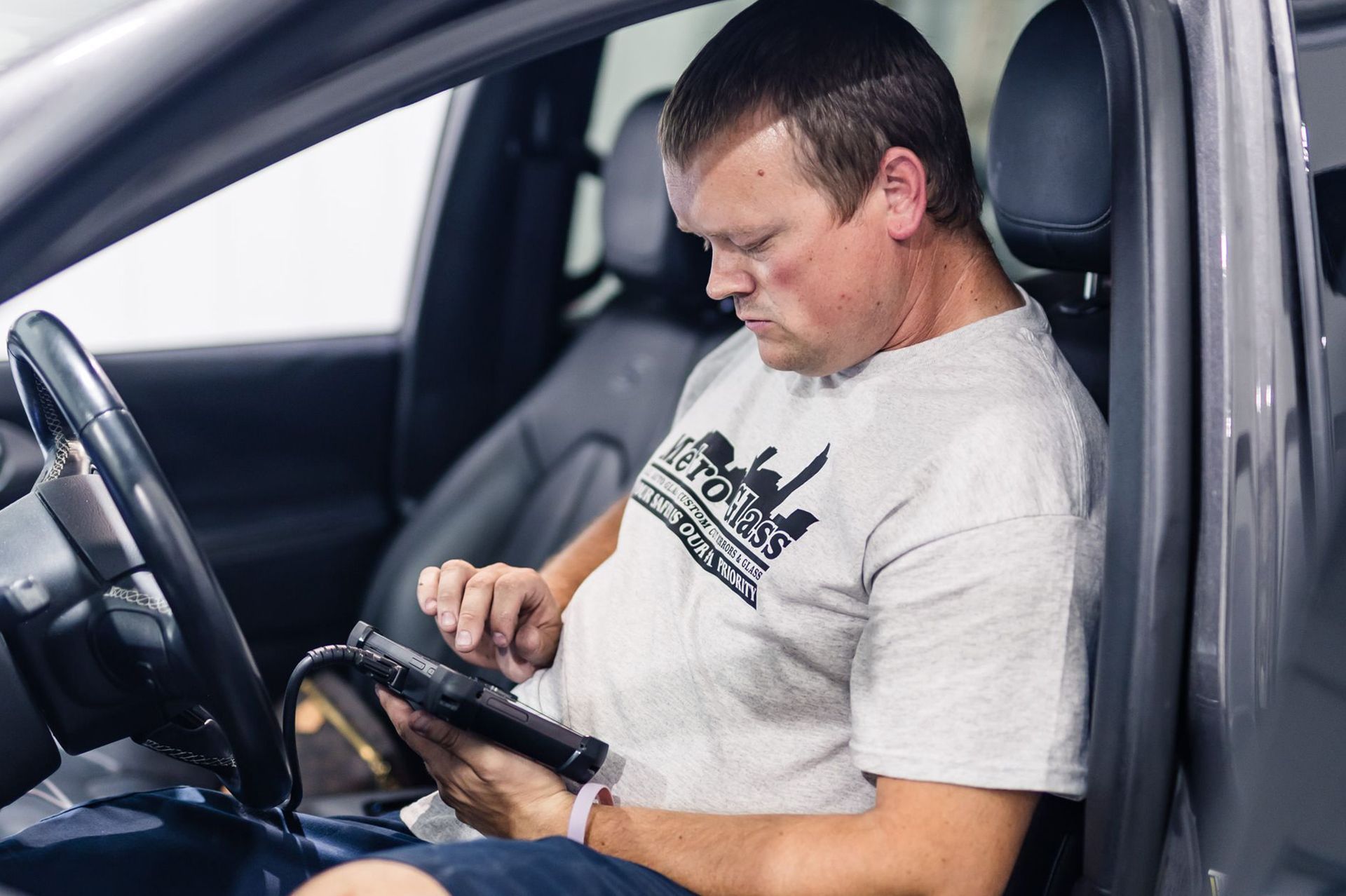 A man in a car using a diagnostic tool. He is wearing a grey t-shirt and sitting in the driver's seat.