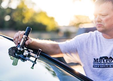 Man repairing windshield outdoors with repair tools, wearing a white Metro T-shirt in sunlight.
