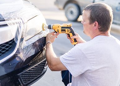 A man using a drill with a buffing pad on a car bumper, outdoors.
