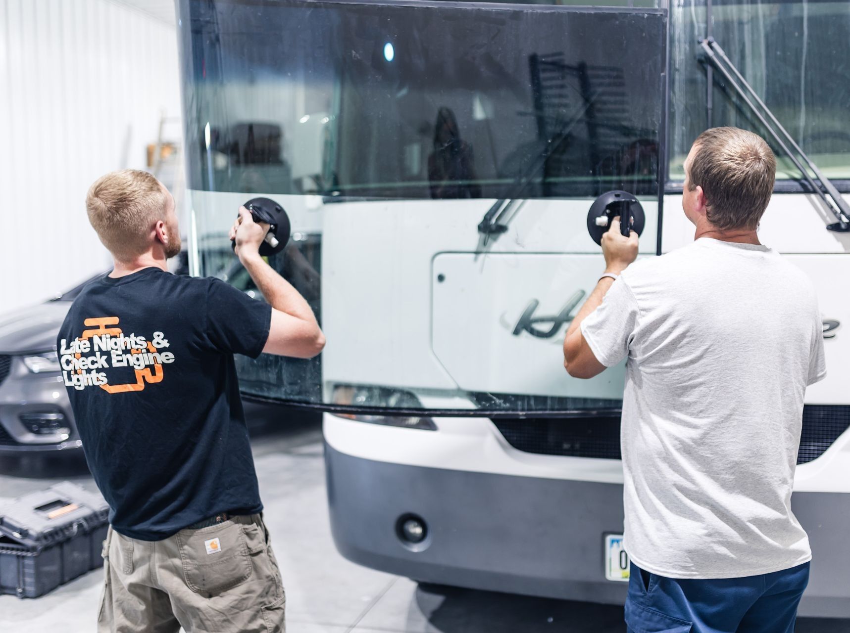 Two men replacing a windshield on a white RV inside a garage.