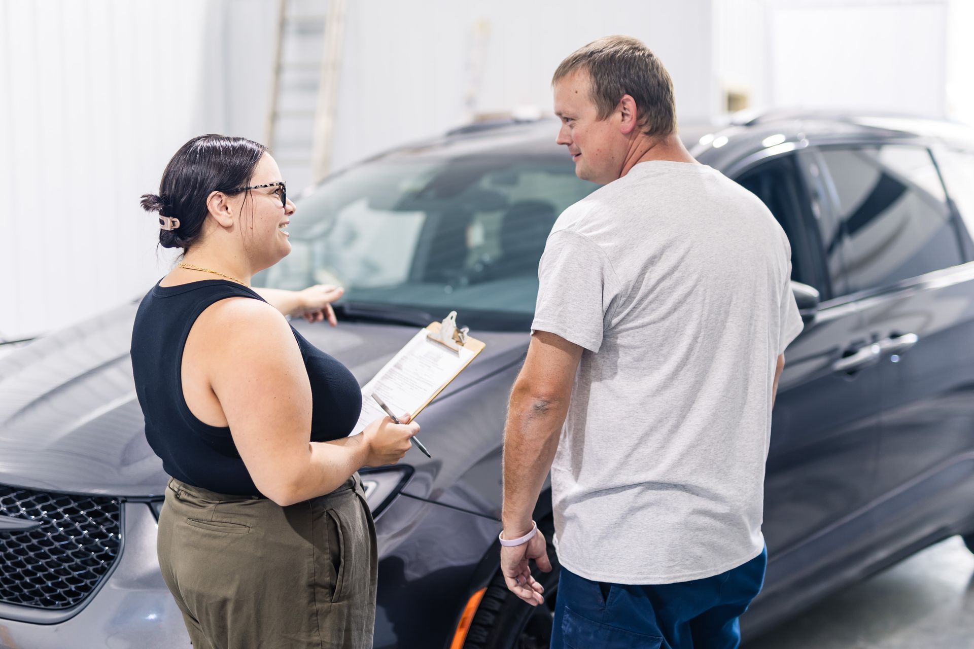 A woman and a man are discussing a car's windshield. The woman holds a clipboard, pointing, while the man looks on. They're in a garage.
