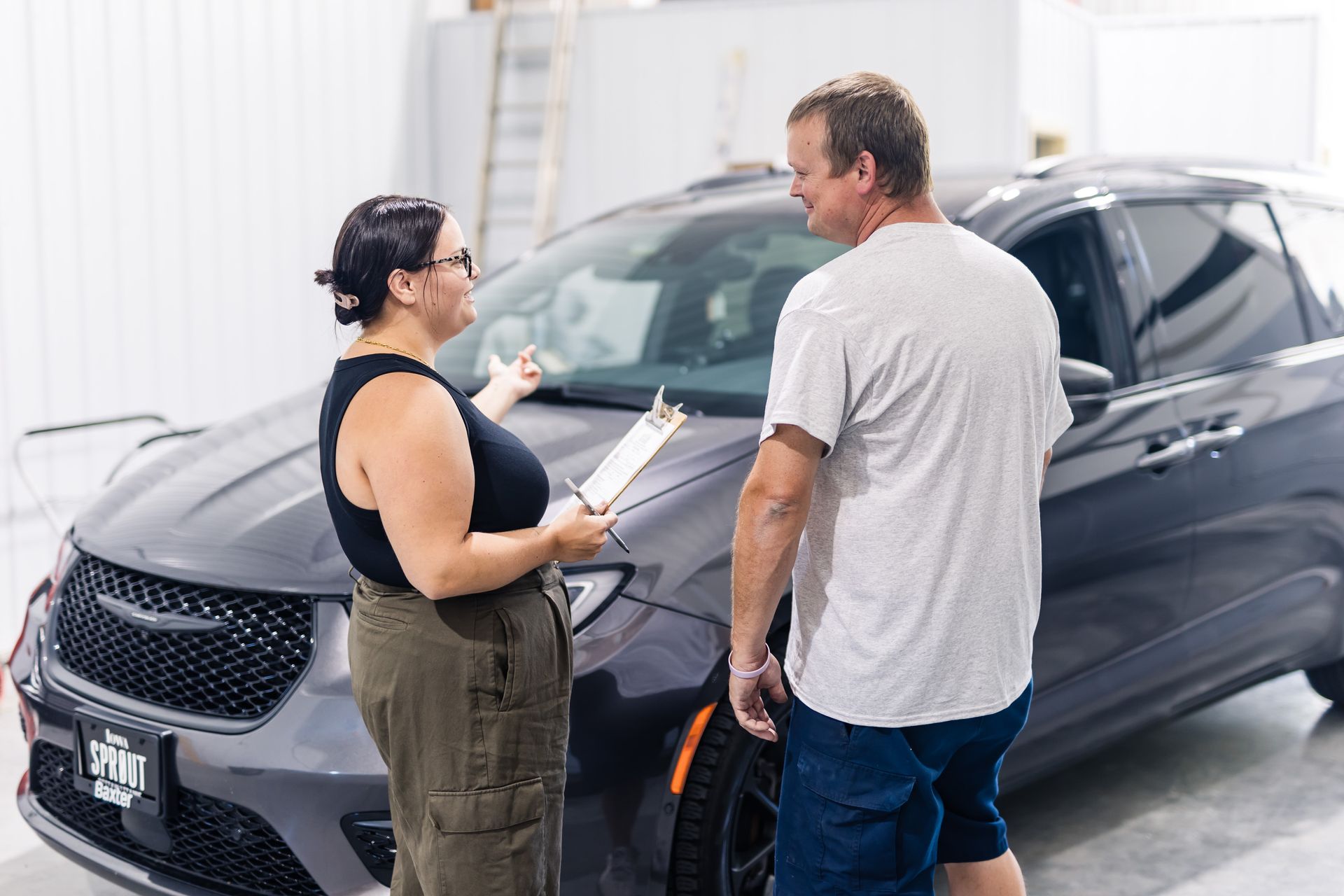 Woman showing a man a clipboard next to a gray minivan in a garage. Both are smiling and talking.