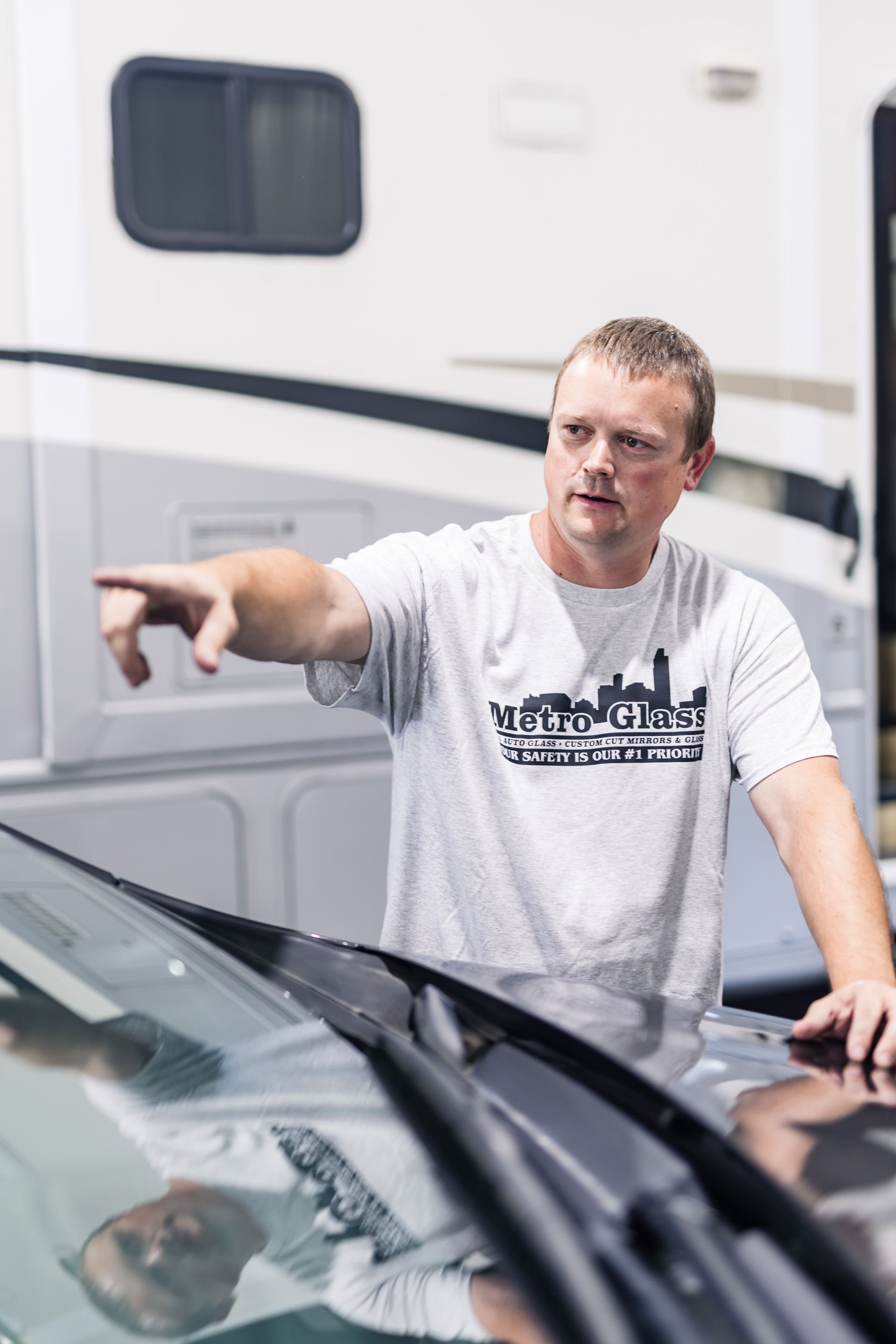 Man pointing at something with his right hand. He wears a light gray t-shirt and stands in front of a car with a large RV in the background.