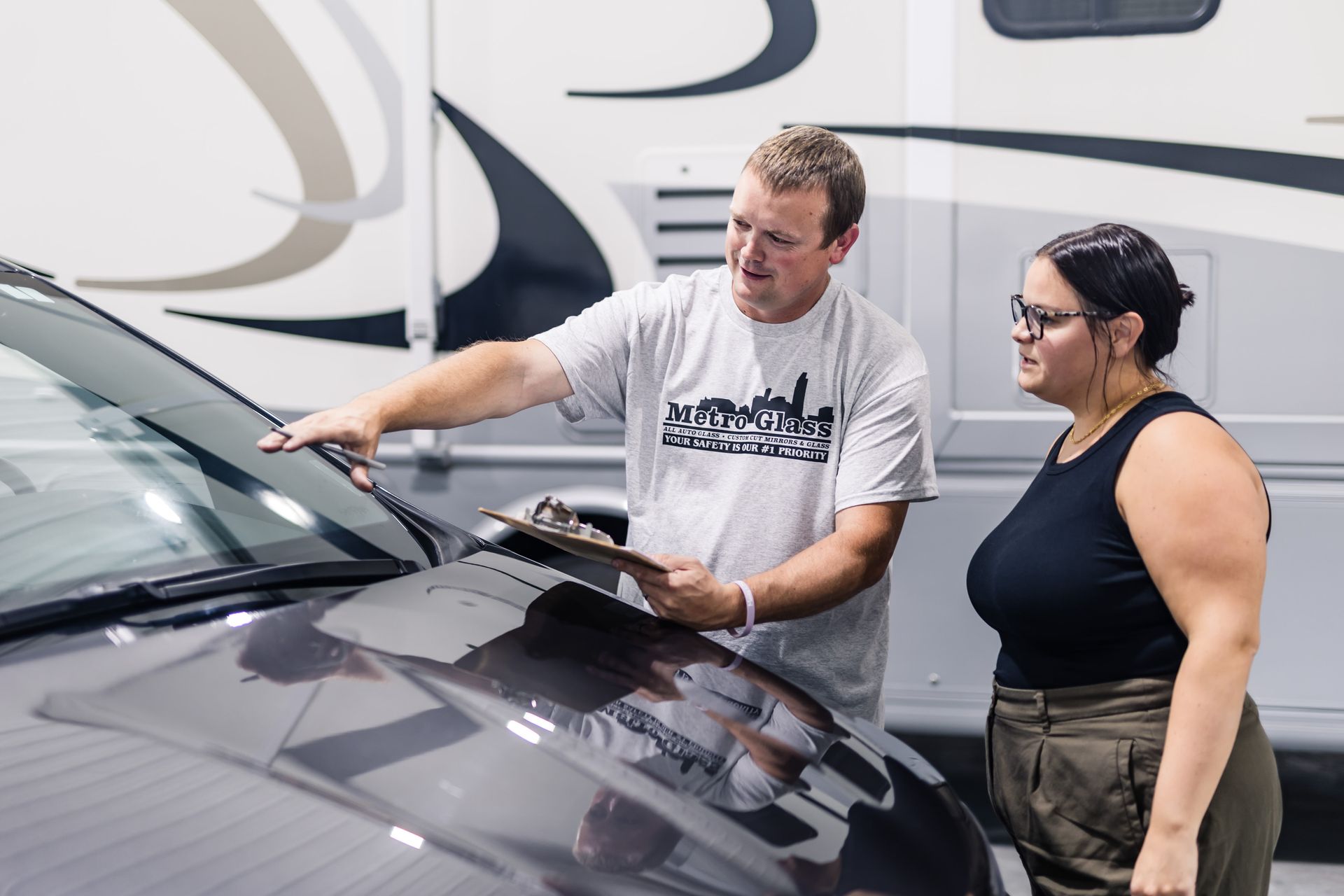 Man pointing at a car windshield, showing a woman a clipboard. They stand near a large RV outdoors.
