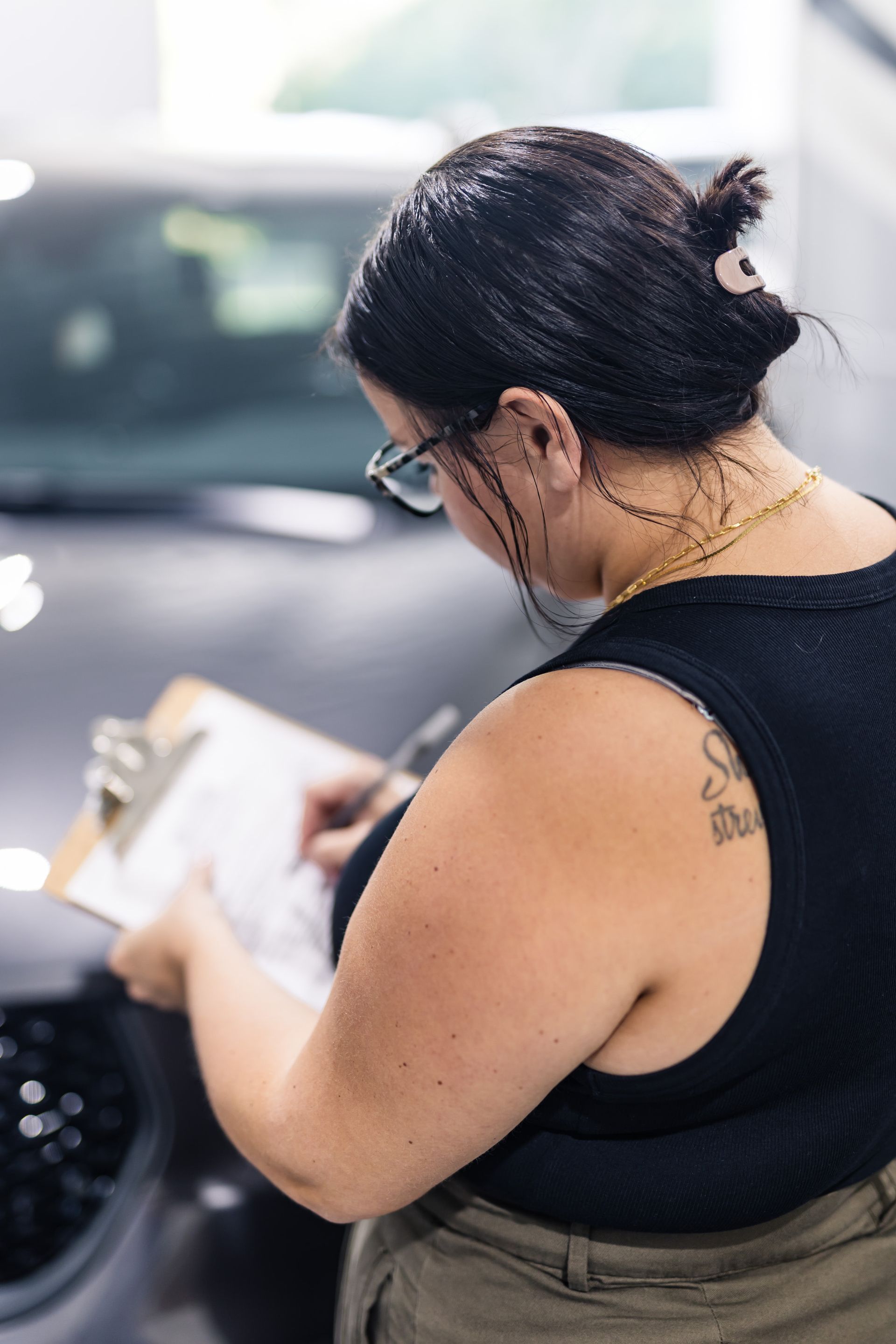 Woman with dark hair, glasses, and a tattoo on her shoulder, writing on a clipboard in front of a car.