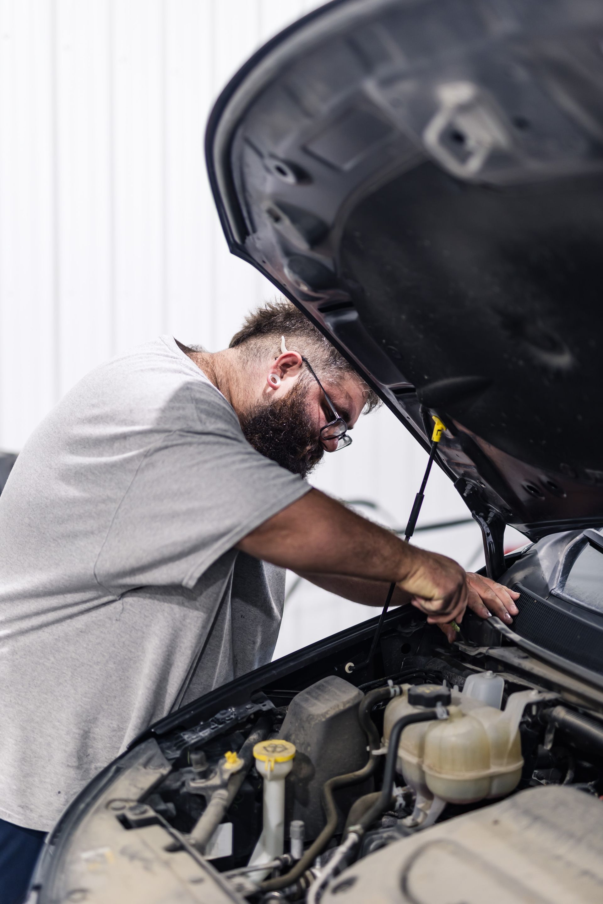 A mechanic with a beard and glasses works on a car engine under the open hood in a garage.
