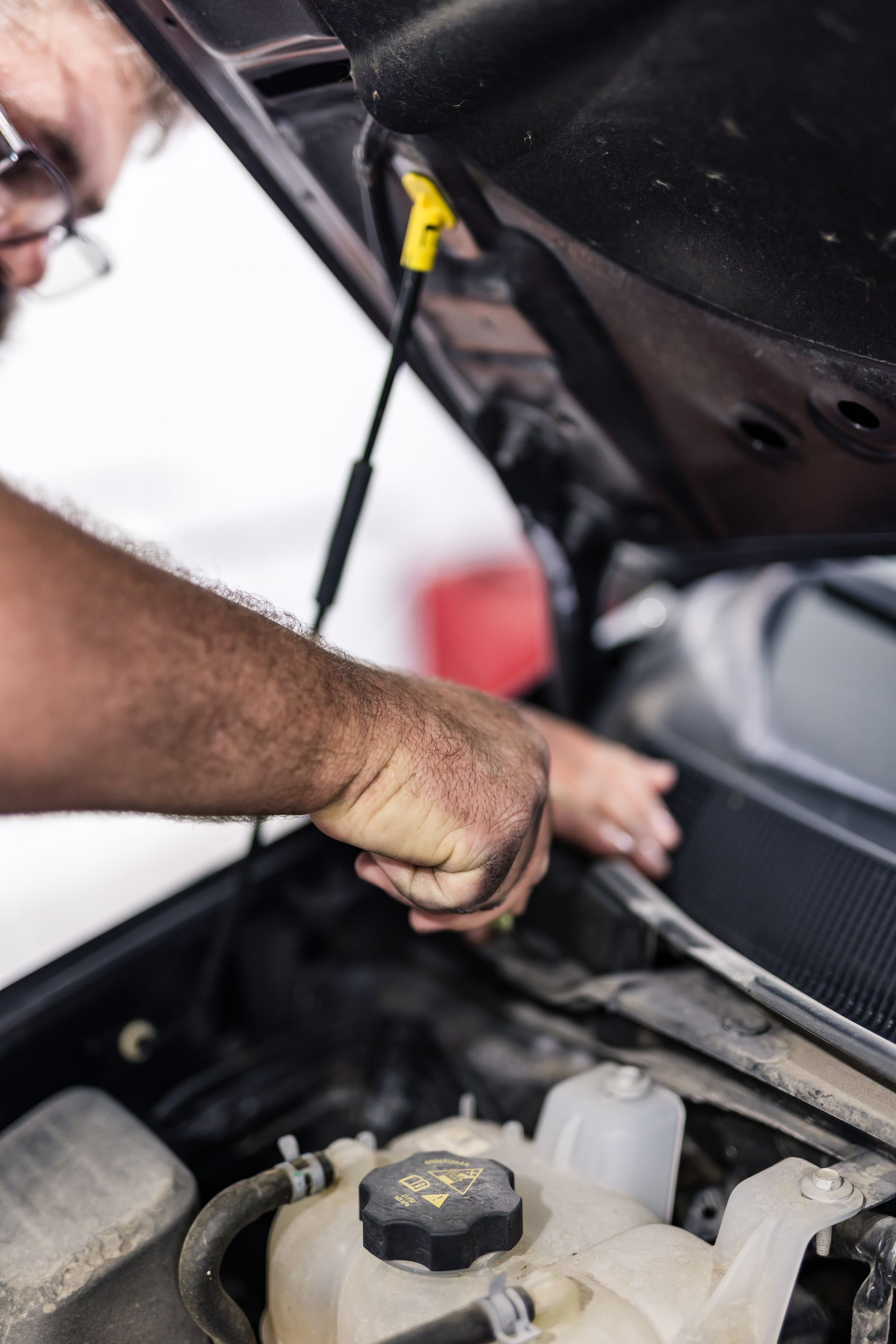 Person with glasses working on a car engine, hood open. Black and yellow components visible.
