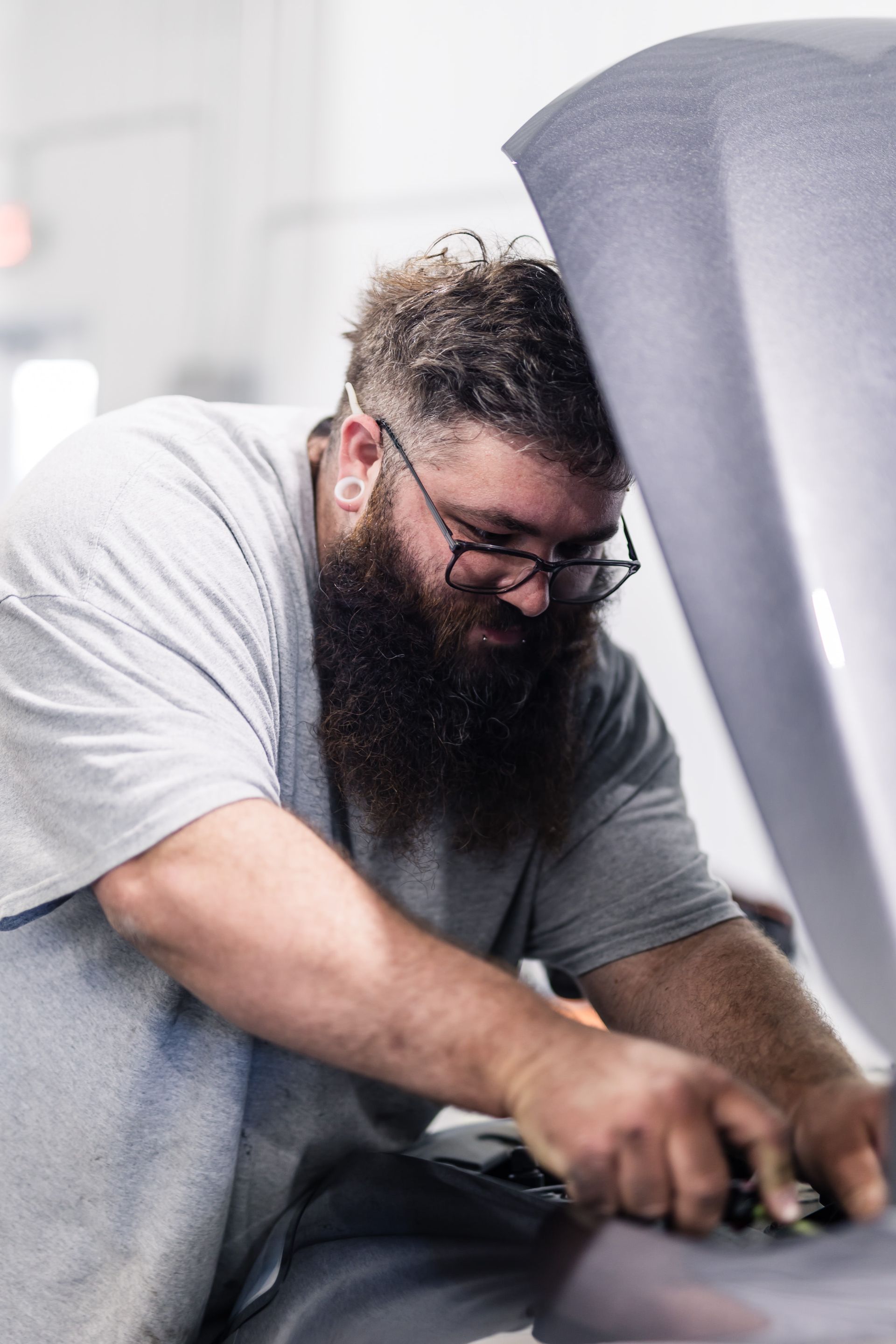A man with a long beard and glasses works on a car, wearing a gray shirt in a garage.