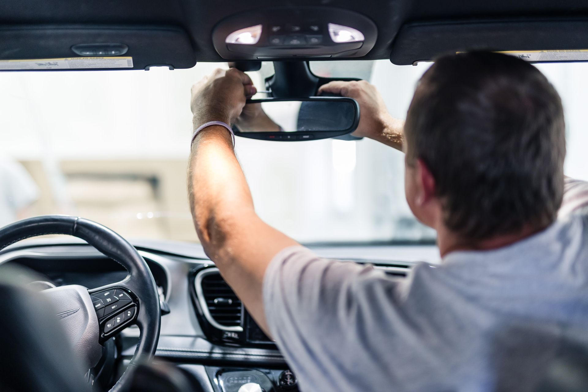 A man adjusting the rearview mirror in a car. He is inside the vehicle, with his hands on the mirror.