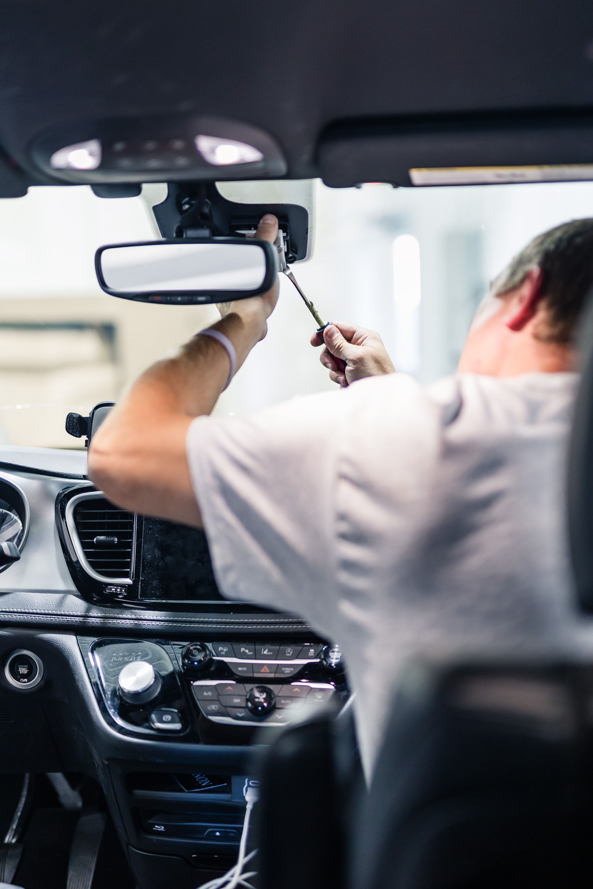 Person working inside a car, likely installing or adjusting something near the rearview mirror. Interior of a modern car is visible.