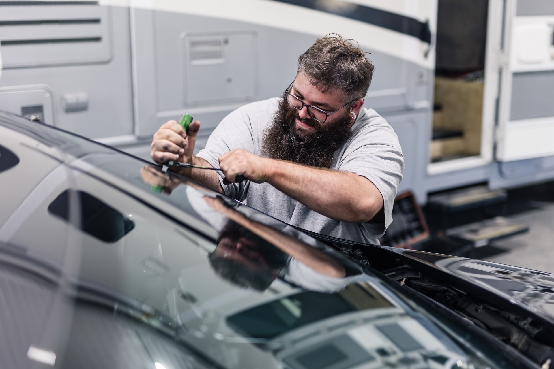 A bearded man in glasses installing something on a car's windshield. He's in a garage, with an RV in the background.