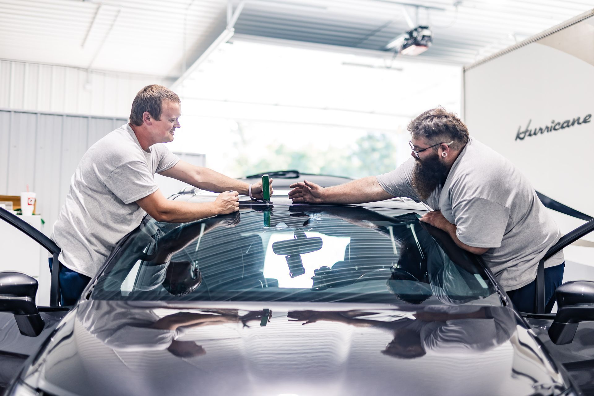 Two men installing tint on a car windshield inside a garage. They wear gray shirts, working together to align and secure the film.