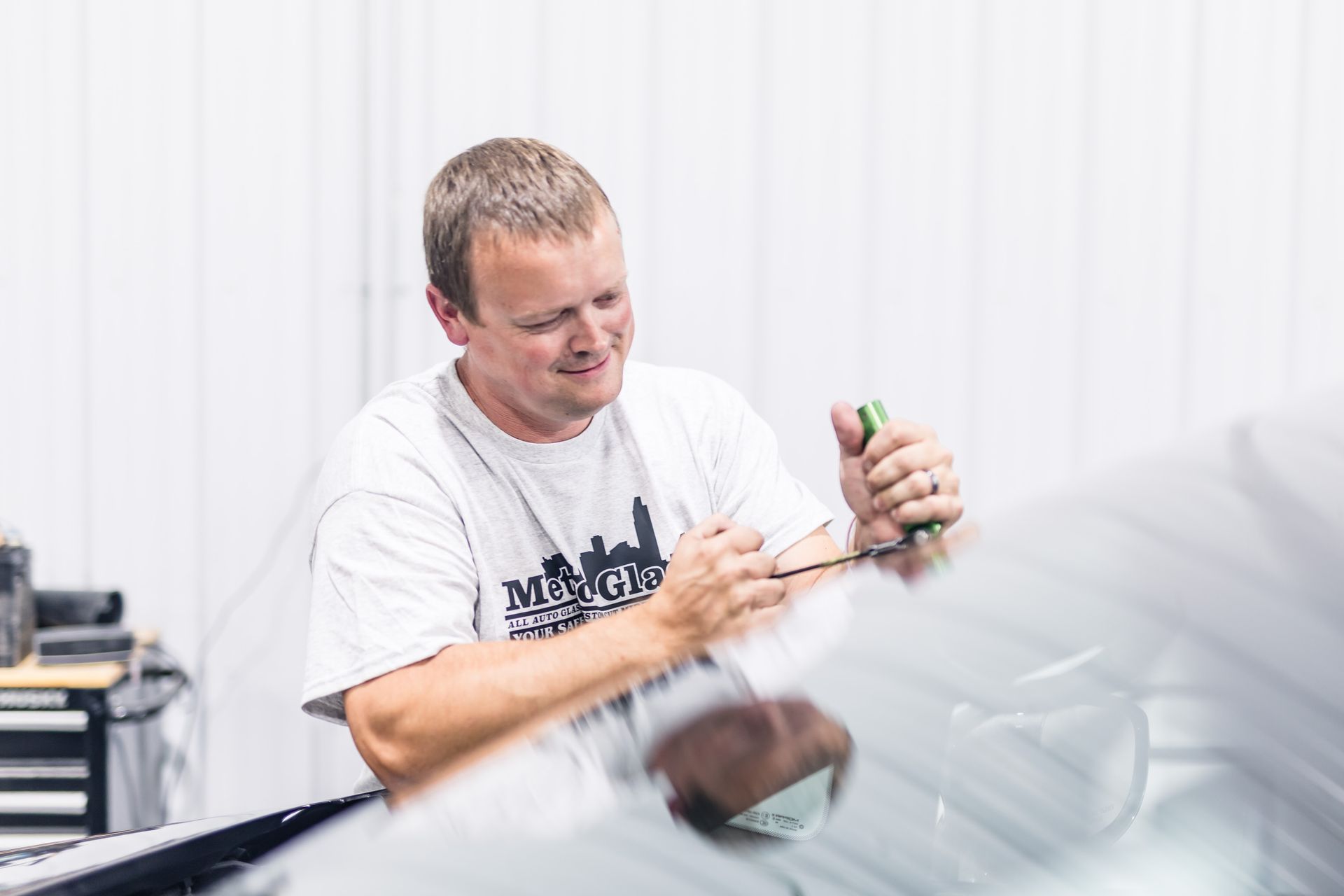 Man applying film to a car windshield in a well-lit garage. He wears a gray t-shirt and holds tools while working intently.