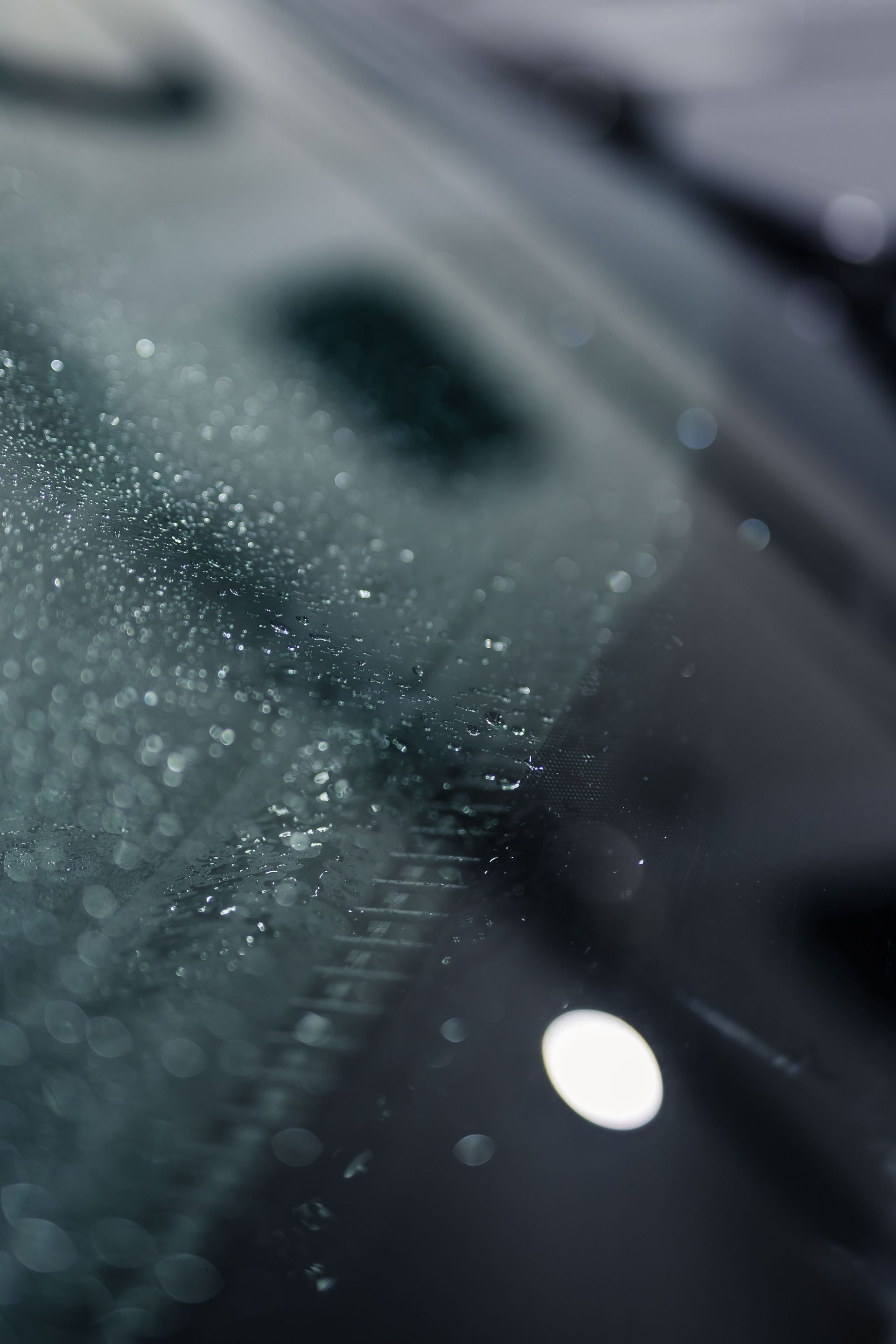 Close-up of a car windshield covered in raindrops. The wipers are partially visible, and a bright, circular light is reflected.