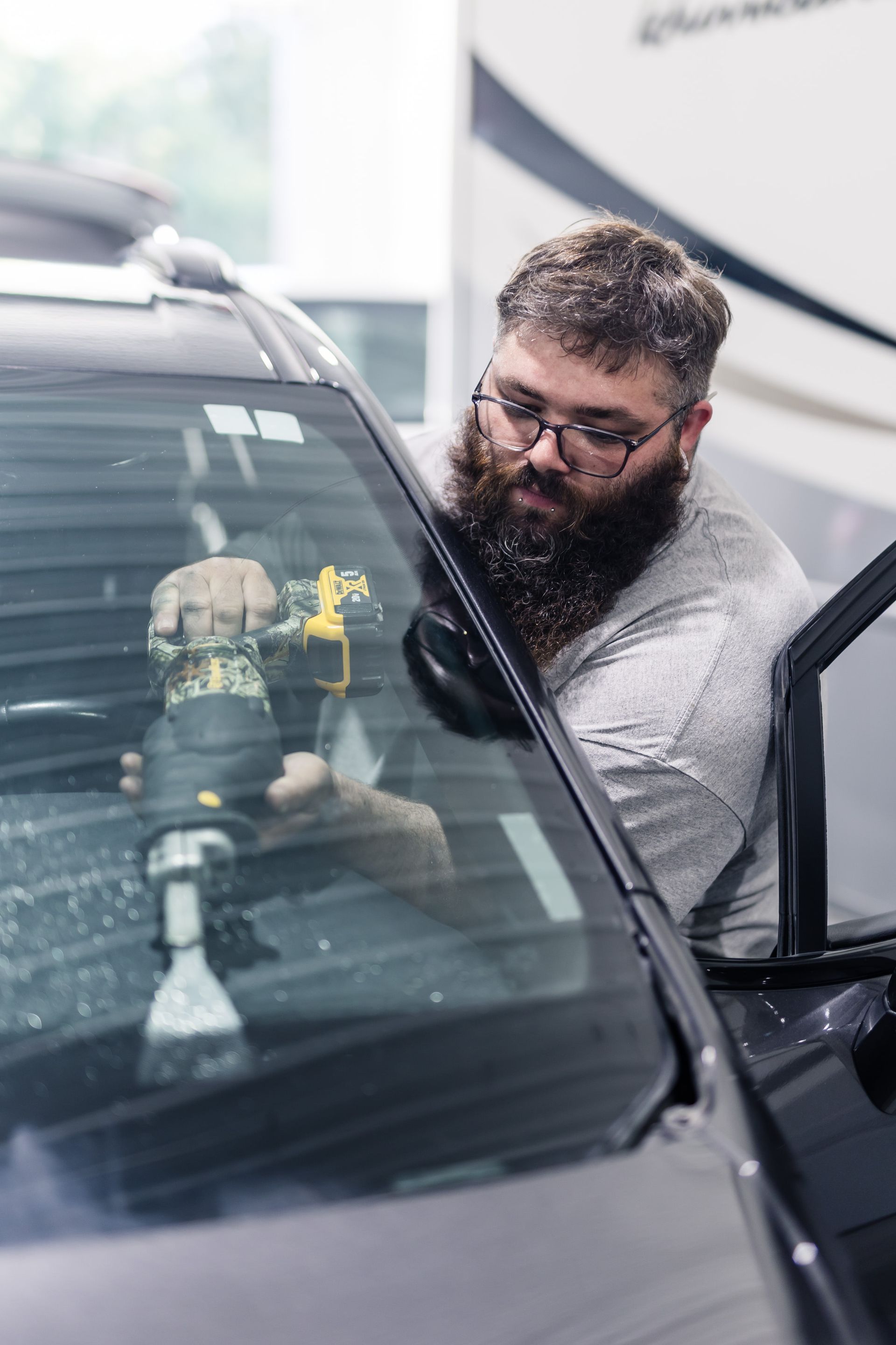 A man with a beard and glasses uses a drill on a car windshield. He's inside the car with the door open in a bright setting.