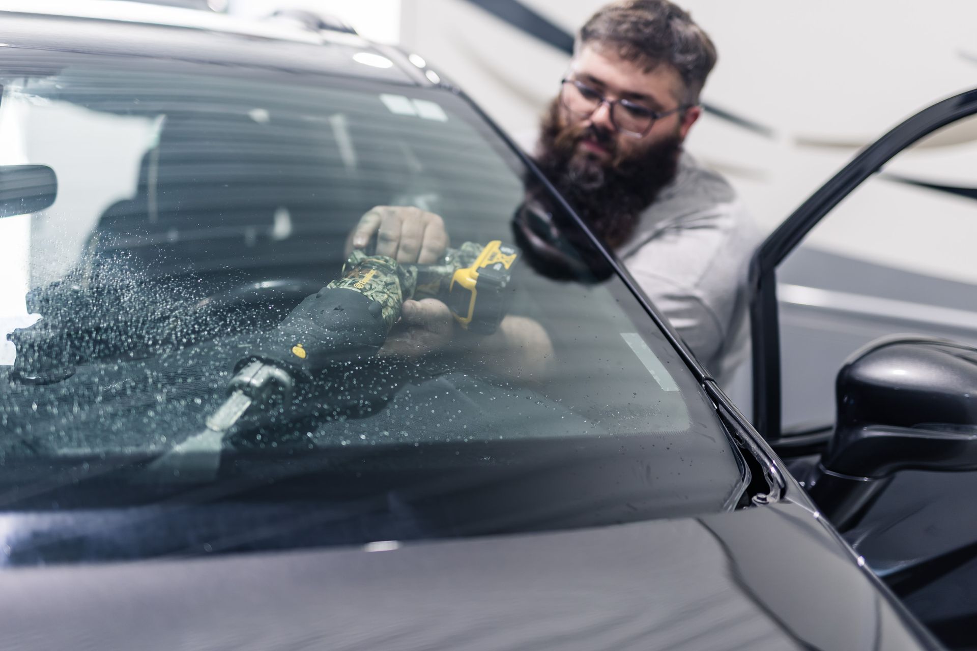 A man with a beard uses a power tool on a car windshield. The car is black and the man is inside with the door open.