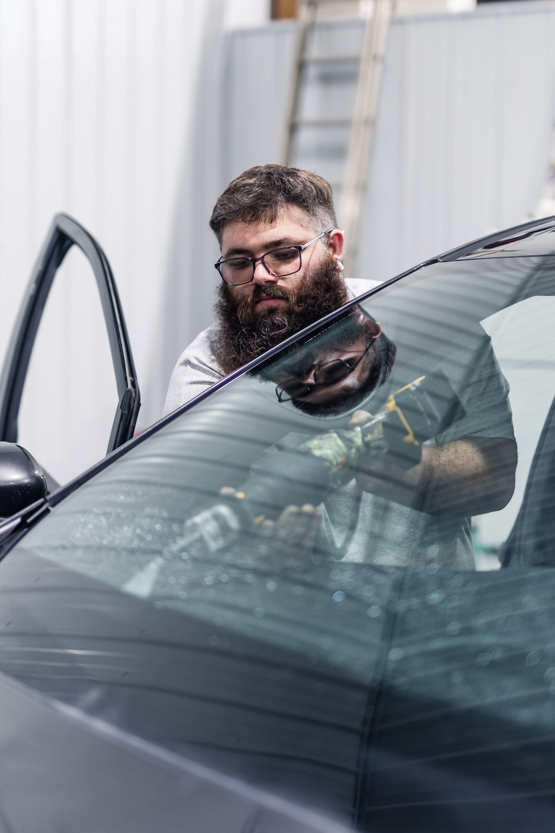 Man with a beard and glasses applying tint to a car's back window inside a shop.