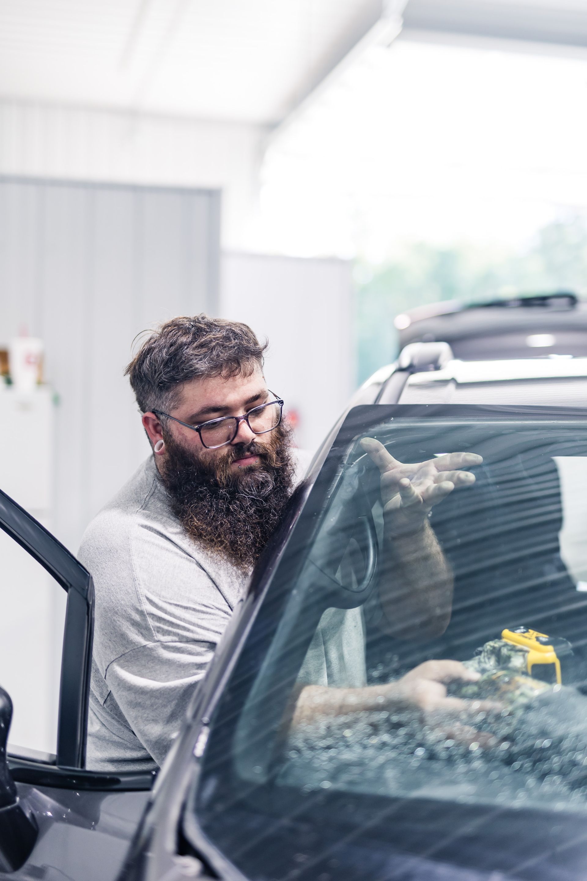 A man with a long beard and glasses applies something to a car windshield in a garage.
