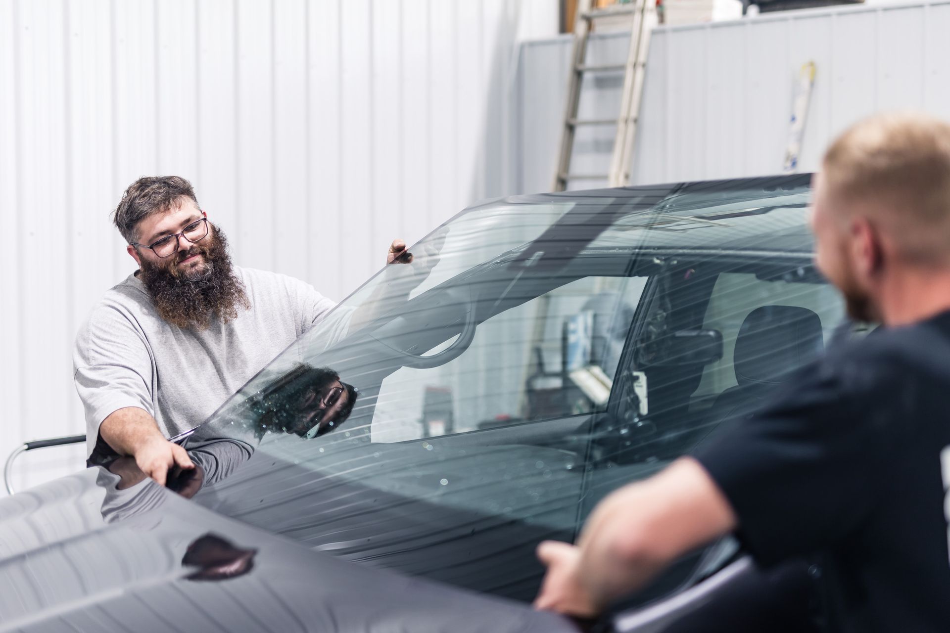 Two men installing a windshield in a garage. One man has a long beard and is smiling while holding the glass.