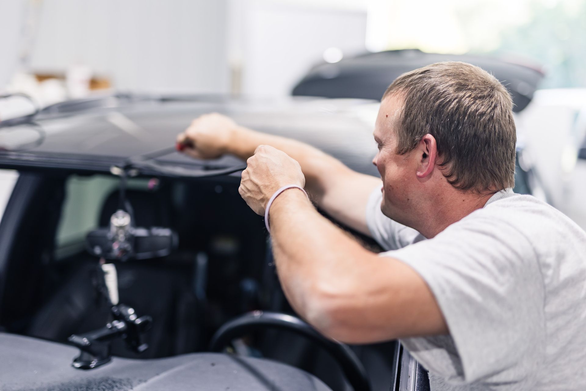 A man installing something on a car's windshield, indoors. He wears a light-colored shirt and focuses on his task.